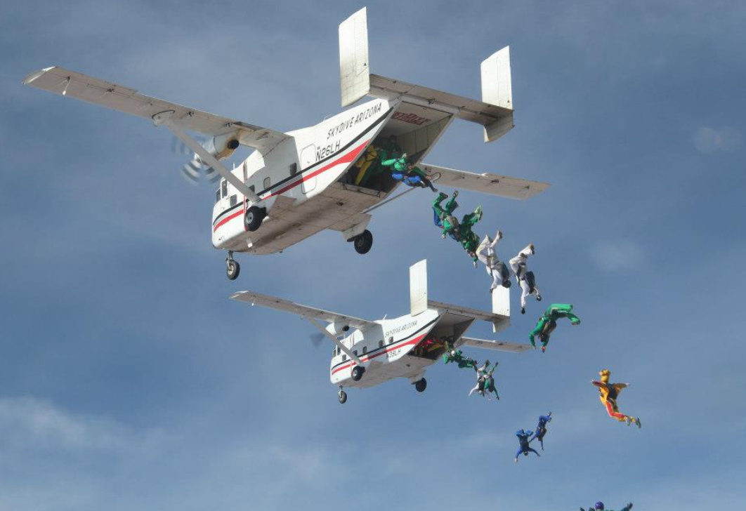 Skydivers exiting two Skydive Arizona Skyvans in colorful jumpsuits during a formation skydive